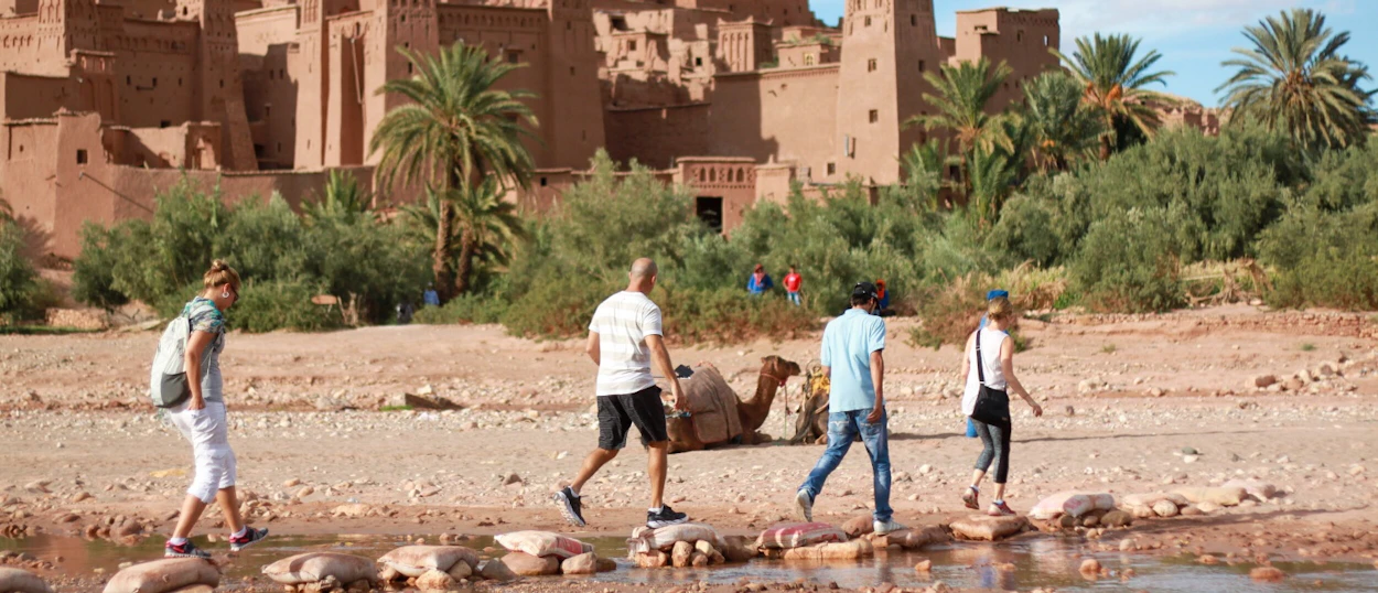 Backpackers cross a shallow river on stepping stones, with the majestic Ait Ben Haddou Kasbah in Morocco.