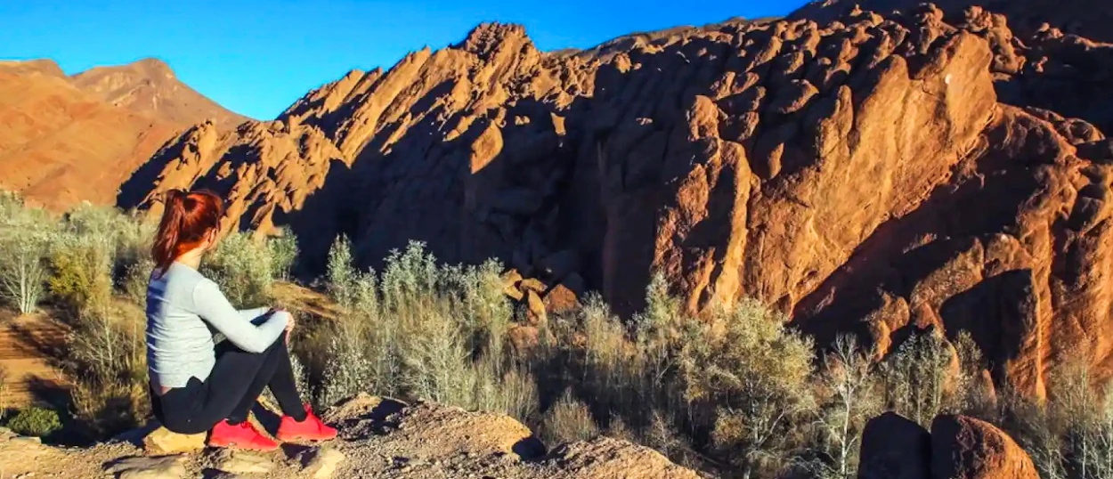 A woman sits on a rock, gazing at the unique reddish rock formations of Dades Valley, Morocco.