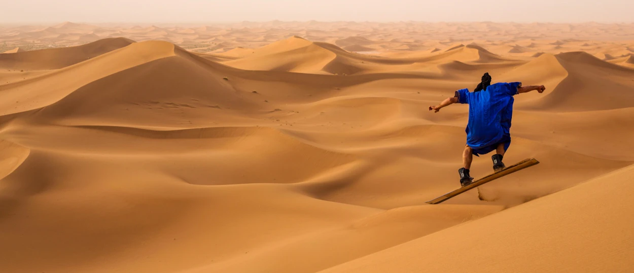An adventurous person in a blue robe sandboards down a steep golden dune in the Merzouga Desert.