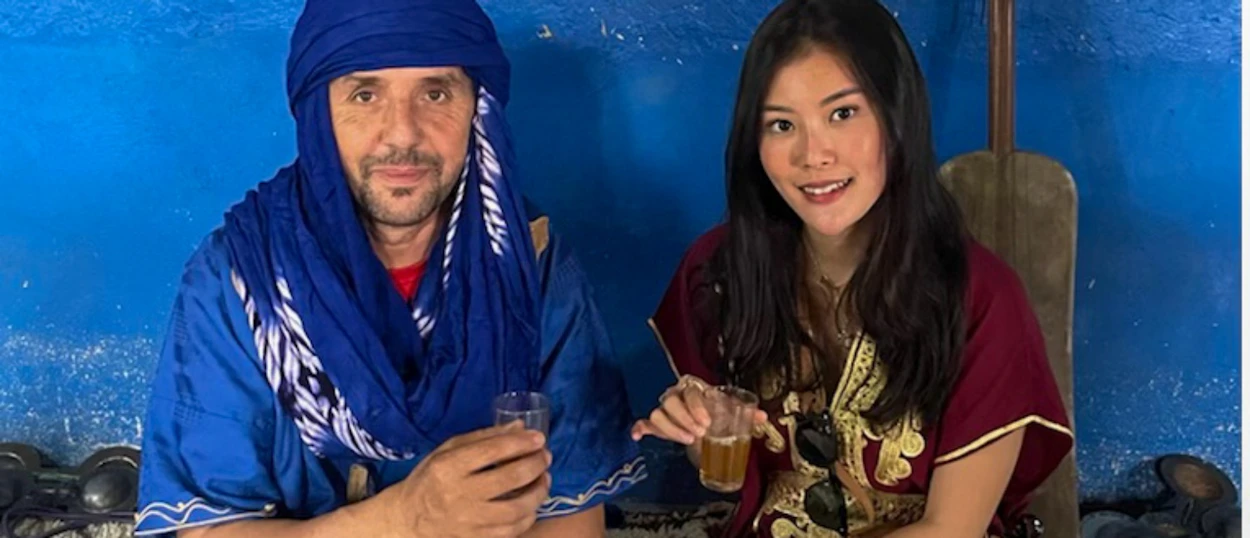 A man in a blue turban and a woman in a maroon tunic share tea, smiling in Morocco.