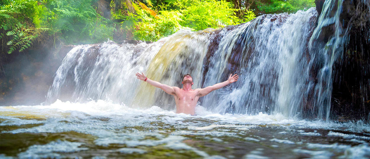 A man stands with arms outstretched under a warm waterfall at Kerosene Creek, Rotorua.