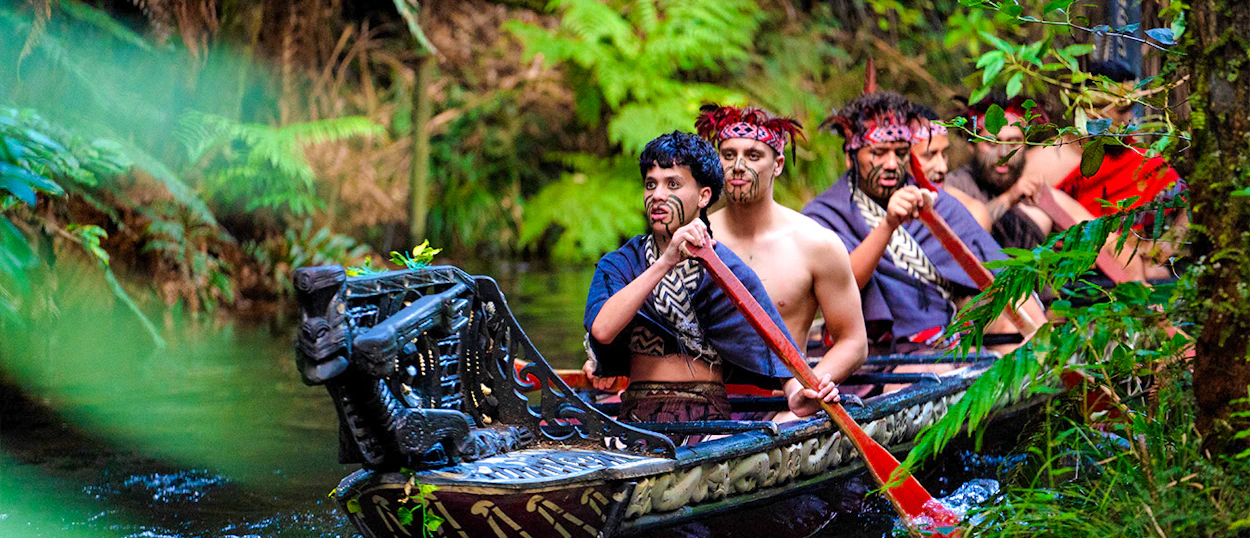 Māori men in traditional dress paddle a waka through a lush forest in Rotorua.