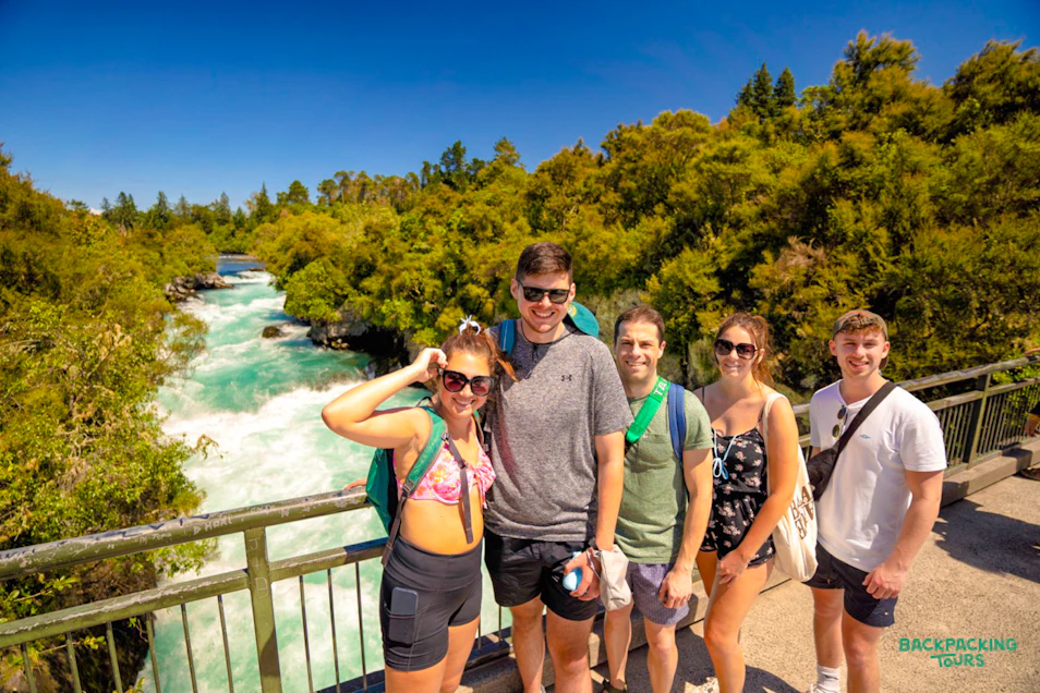 Five happy backpackers pose on a bridge overlooking the stunning turquoise Huka Falls in New Zealand.