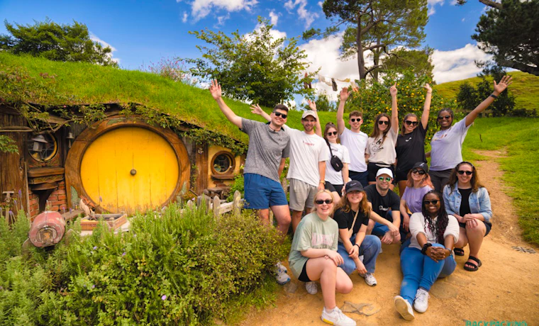 A group of happy backpackers pose in front of a hobbit hole at the Hobbiton film set.