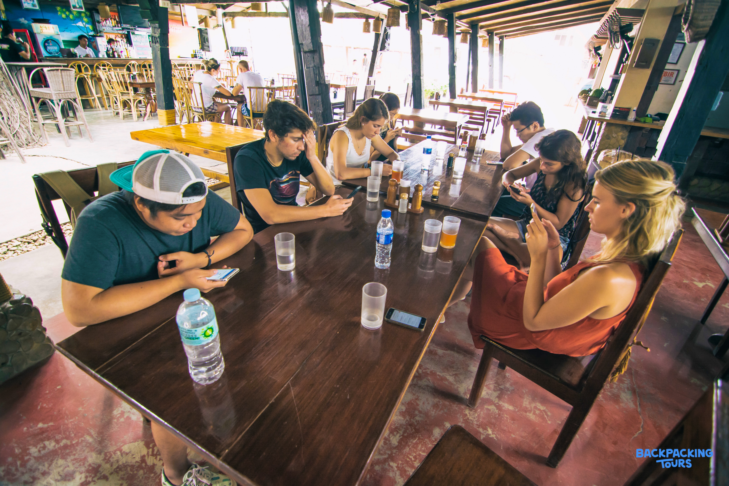 Young travelers relax together in a bright, open-air common area, checking their phones.