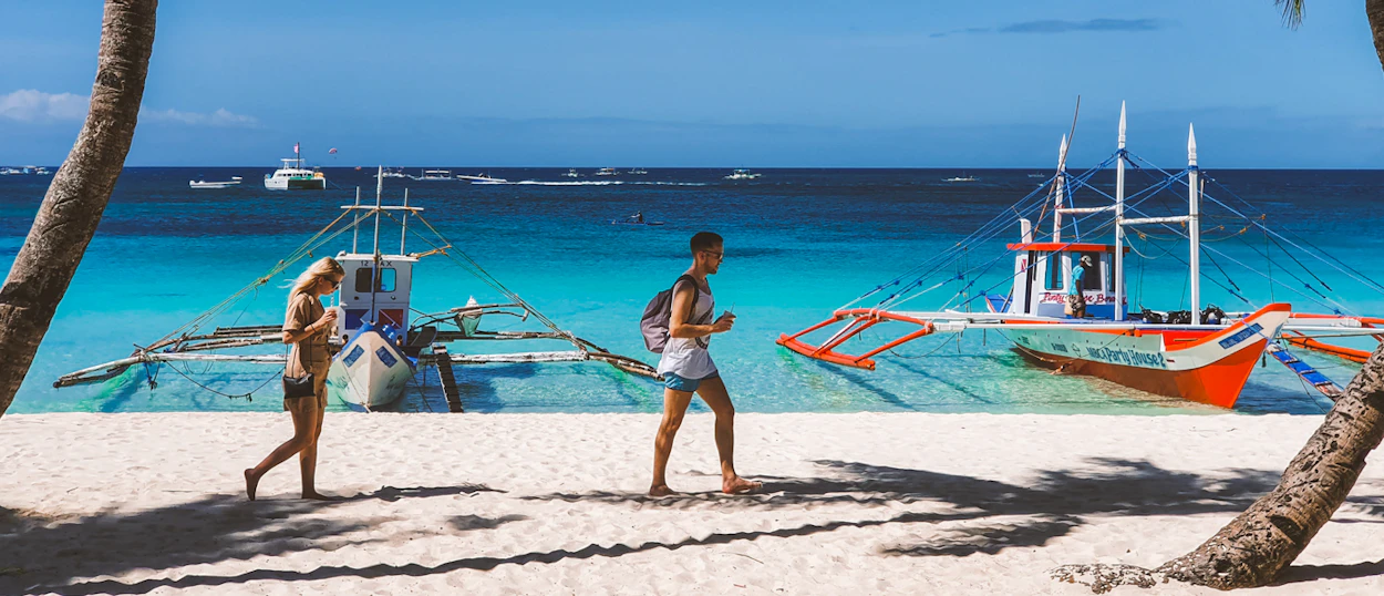 Travelers walk on a white sand beach with turquoise water and traditional outrigger boats in the Philippines.