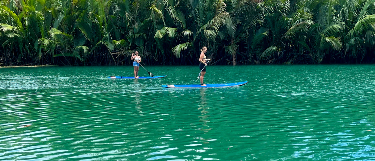 Two people paddleboarding on the vibrant green Loboc River surrounded by lush tropical trees.