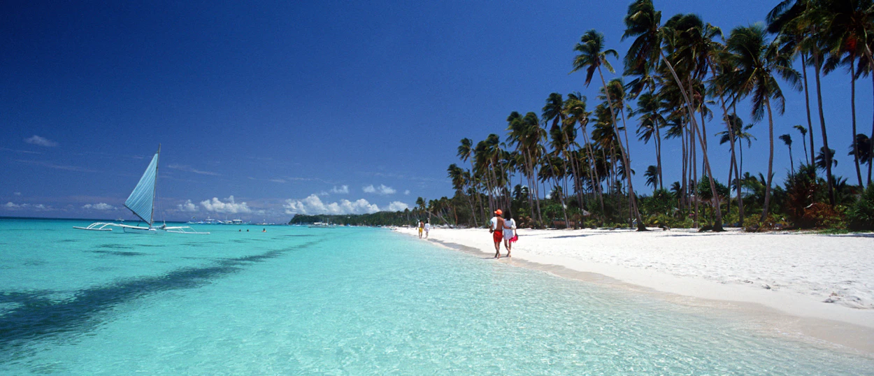 Two people walk on a pristine white sand beach with clear turquoise water and palm trees in Boracay.