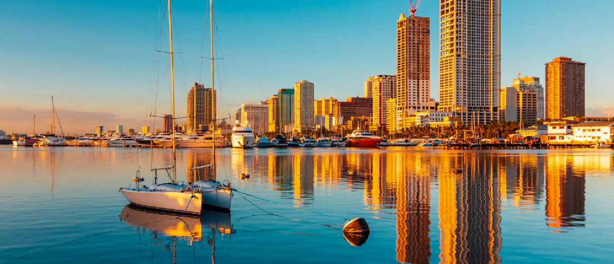 Golden light illuminates Manila's modern skyline and yachts reflecting in the calm bay at sunset.