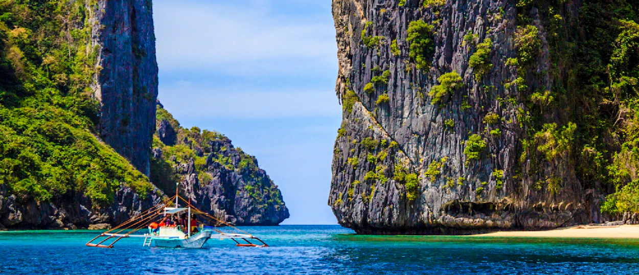 A traditional bangka boat sails past dramatic limestone cliffs and a pristine beach in Coron.