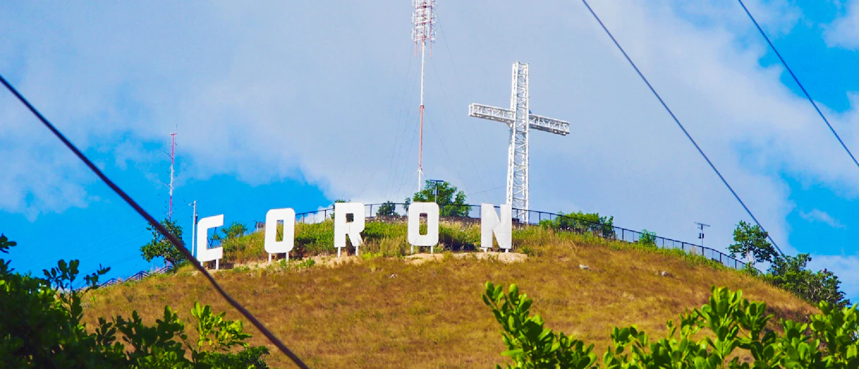Large white 'CORON' sign and a cross on a dry hillside under a blue sky.