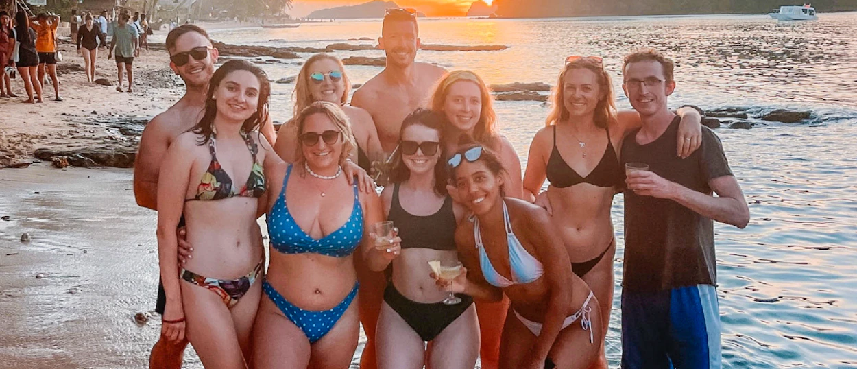 A diverse group of happy friends in swimwear posing on a beach at sunset, enjoying drinks.