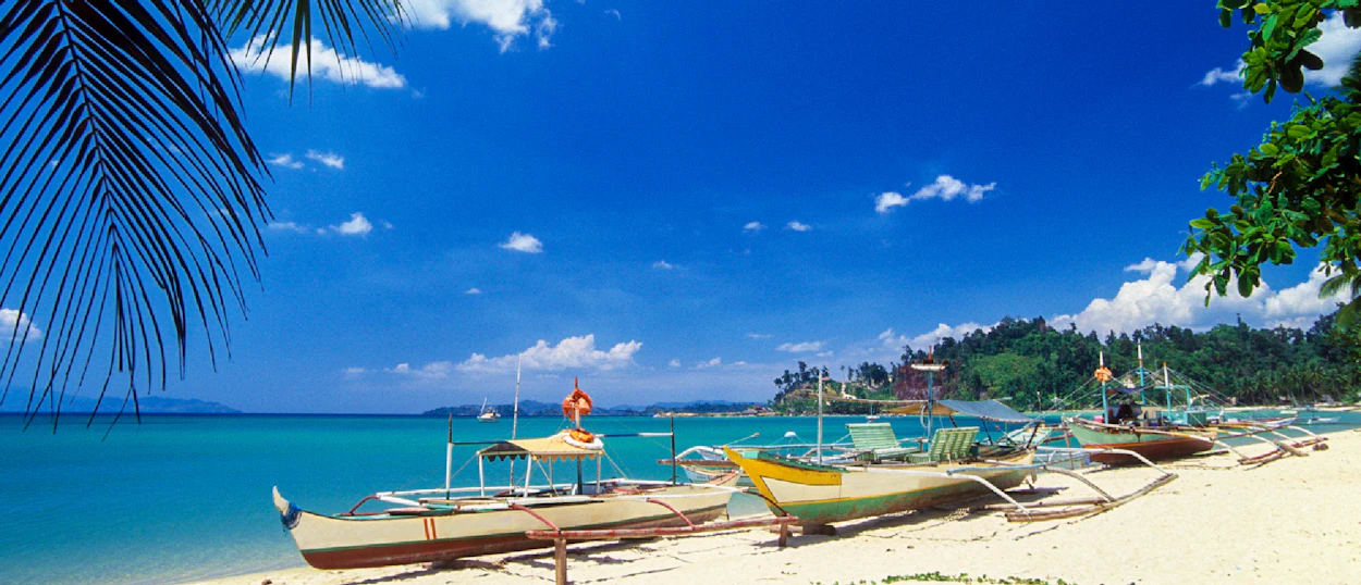 Several traditional boats rest on a white sand beach with clear turquoise water in Coron.