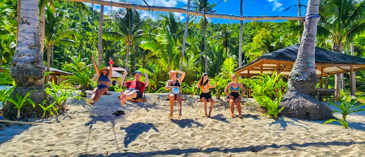 Five happy travelers enjoy a beach swing and sunny sand amidst tropical palms in the Philippines.