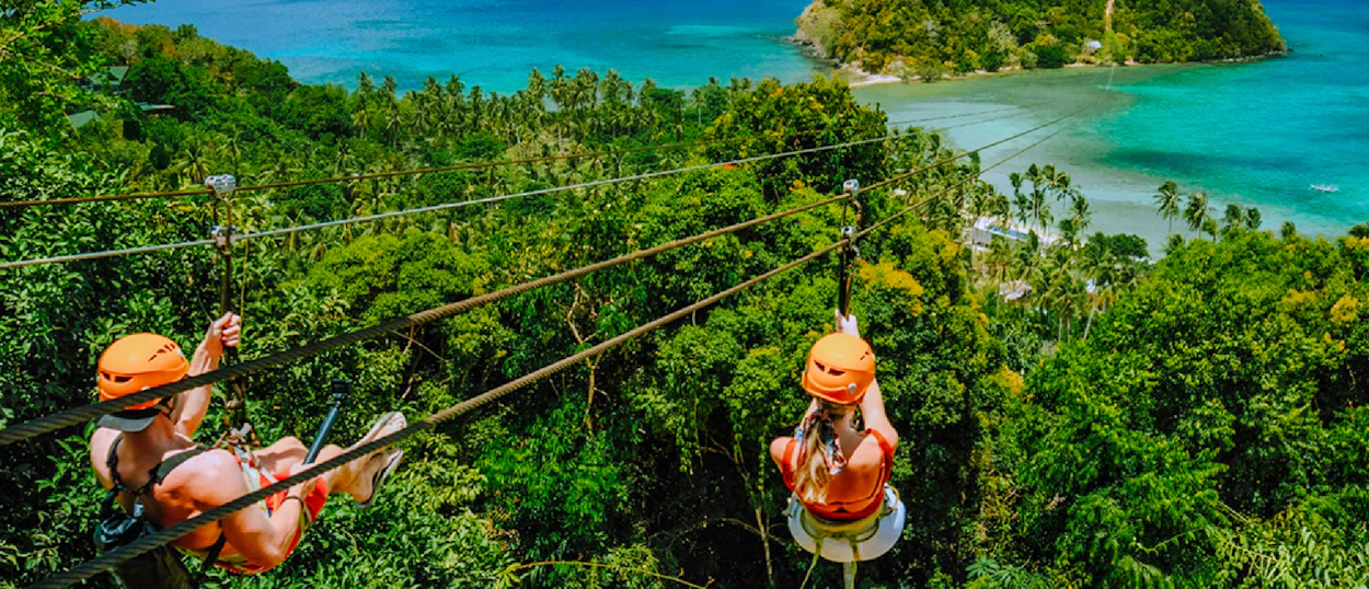 Two people ziplining over a lush green forest with a stunning turquoise bay in El Nido.