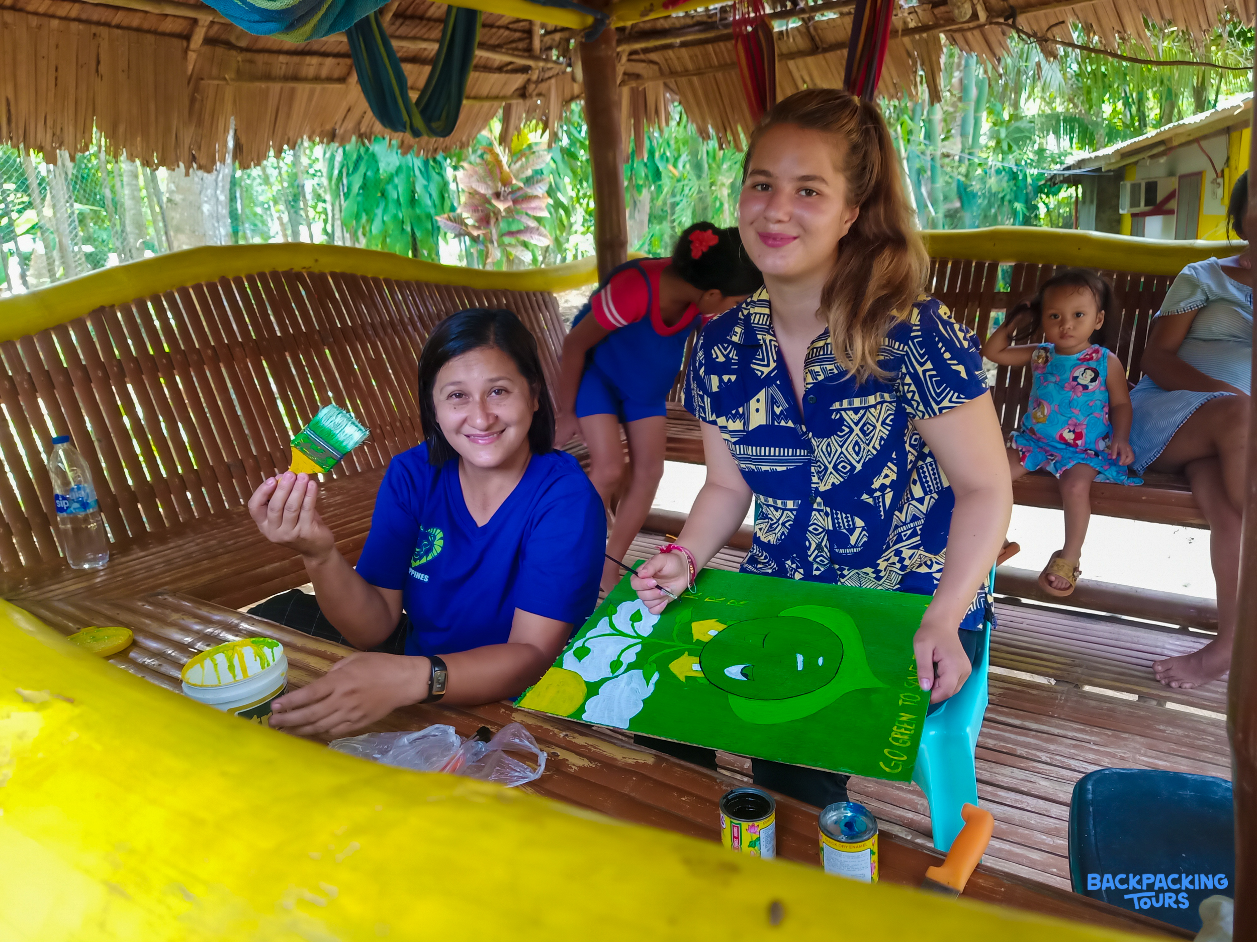 Two women smiling while painting a crocodile on a canvas in a bamboo hut in the Philippines.