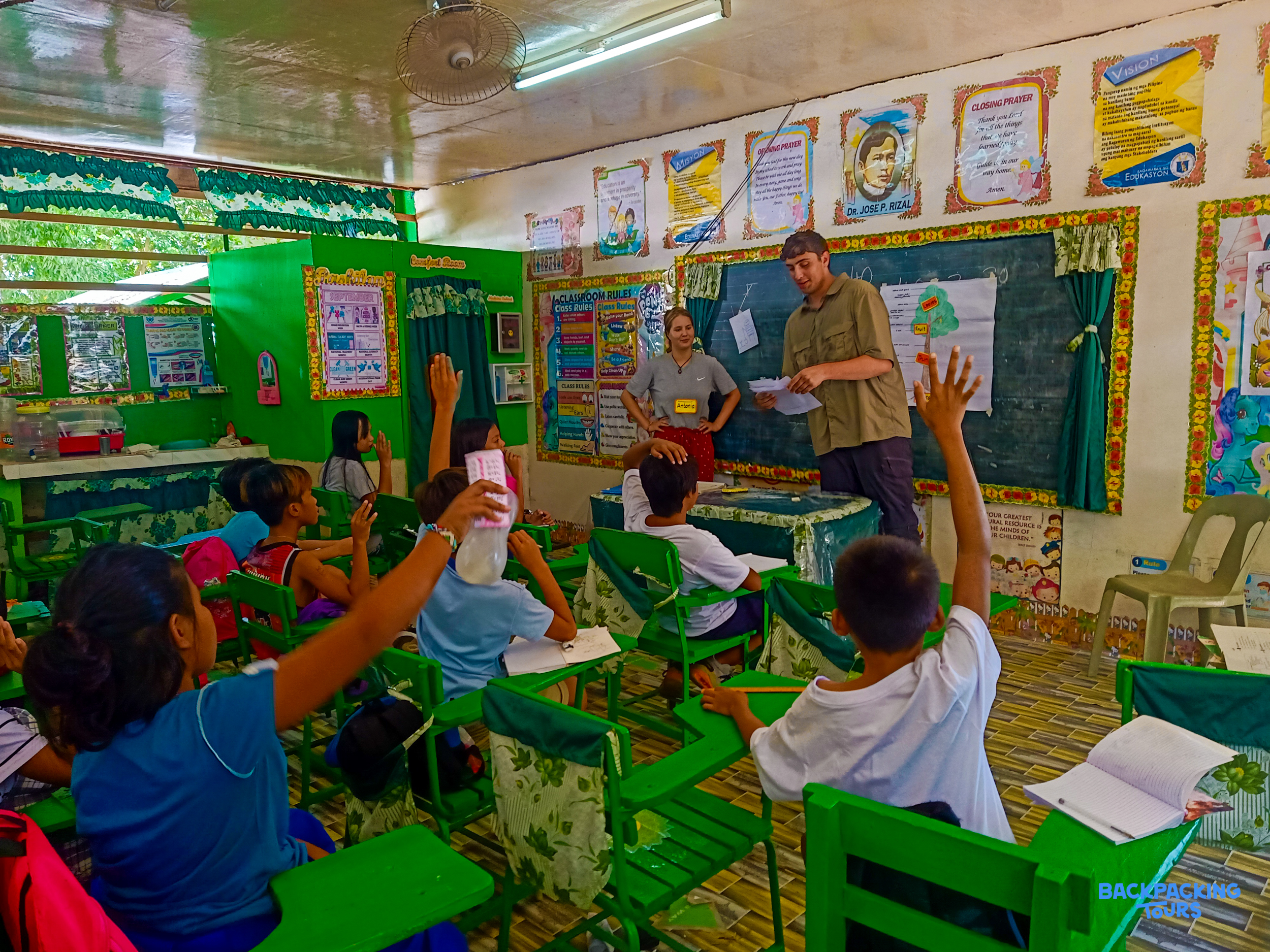 Children in a bright classroom eagerly raise their hands during an interactive lesson led by two adults.