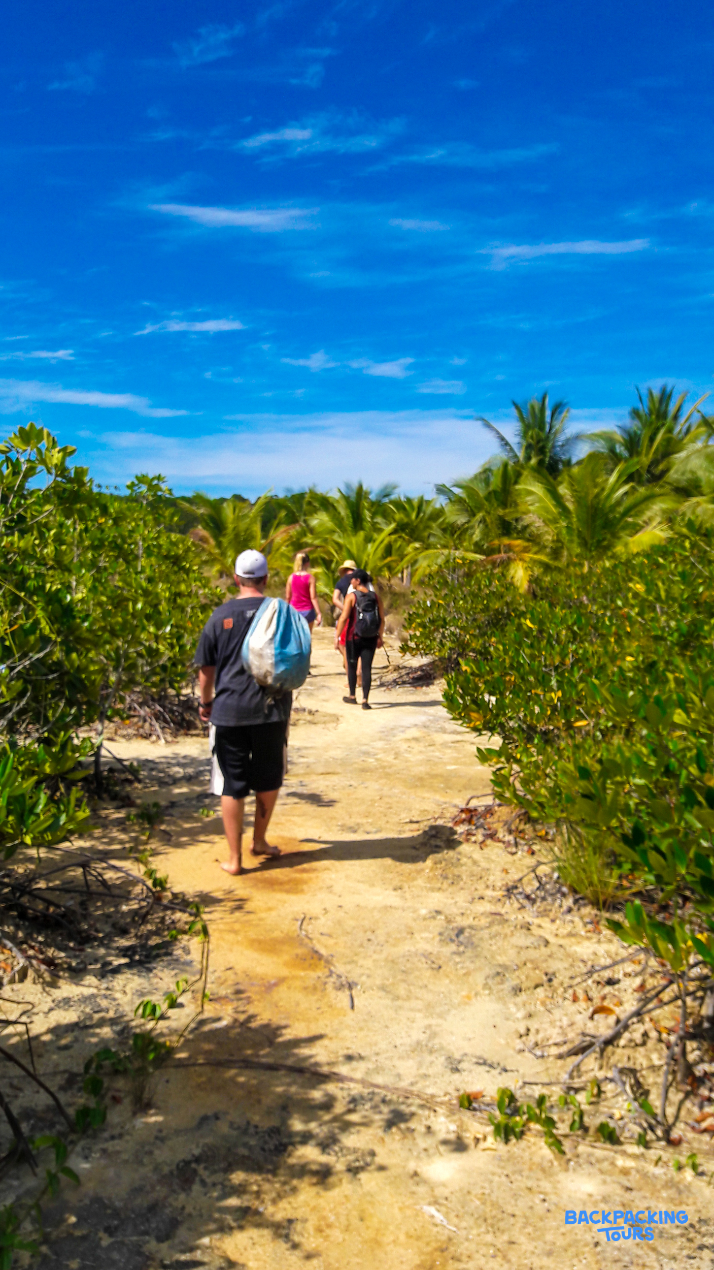 Friends walk a sandy path through lush tropical plants under a bright blue sky in the Philippines.