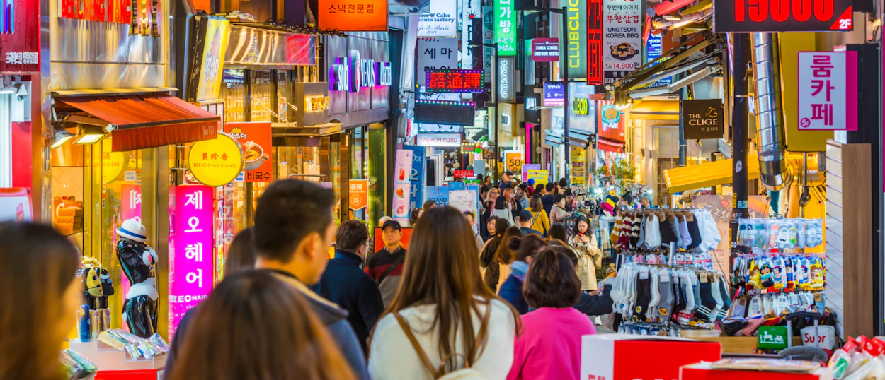 A vibrant, crowded shopping street in Seoul with many people and colorful neon signs.
