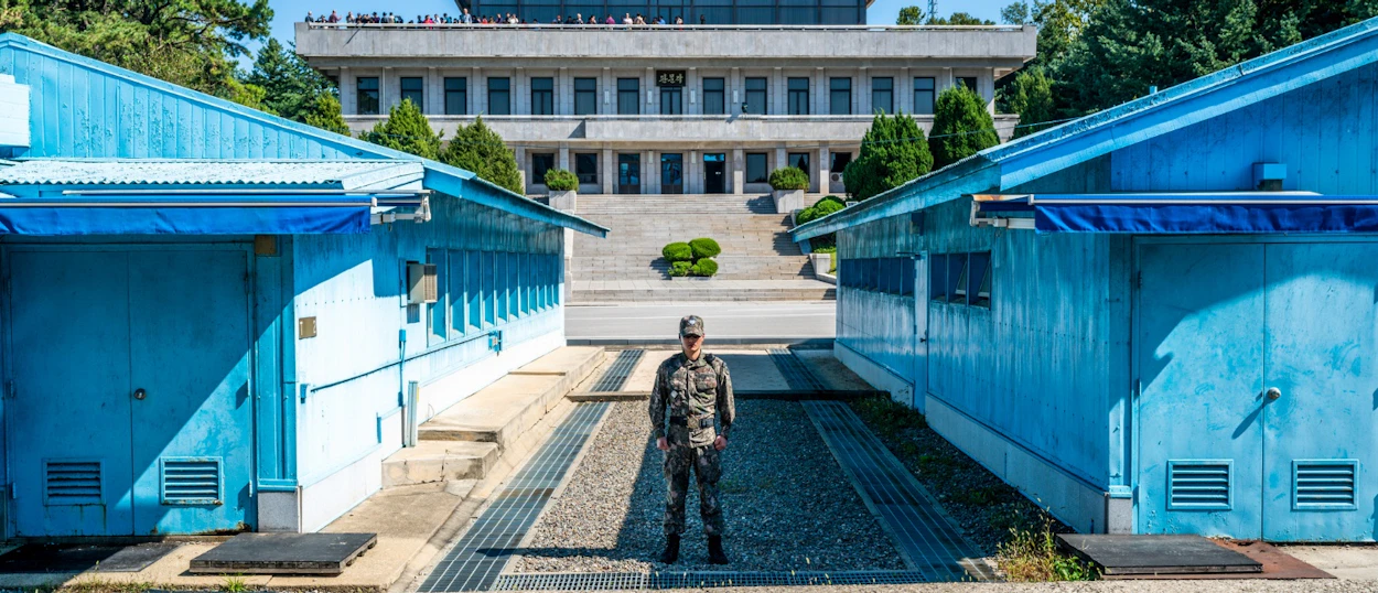 A soldier in camouflage uniform stands between two blue buildings at the Korean Demilitarized Zone.