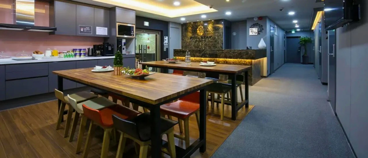 A modern hostel common area featuring a kitchen, long dining tables, and reception desk in Seoul.