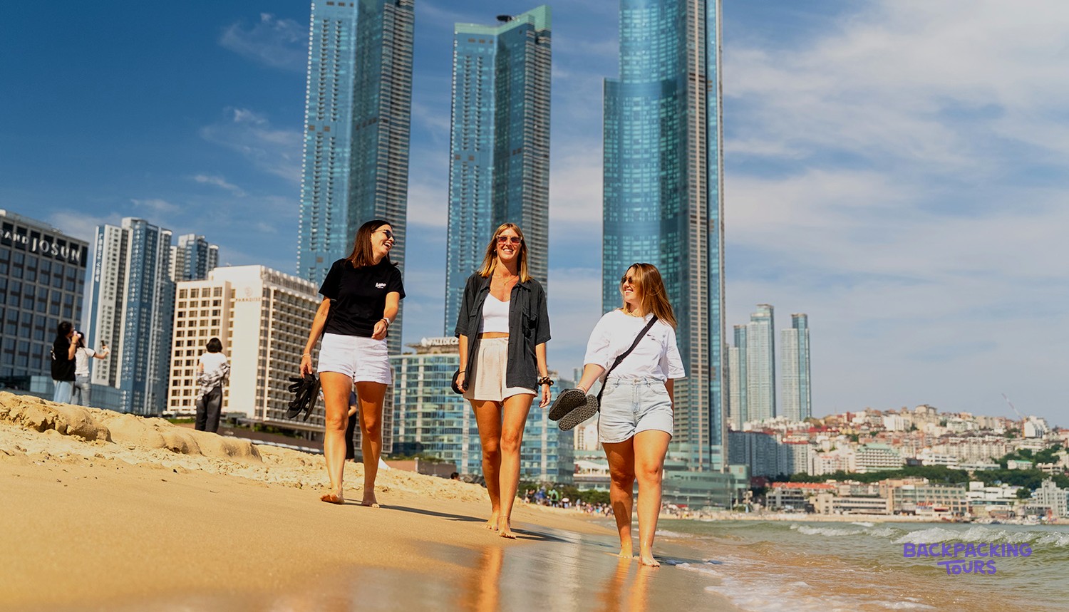 Three travellers walking along Haeundae Beach with the Busan skyline rising behind them.
