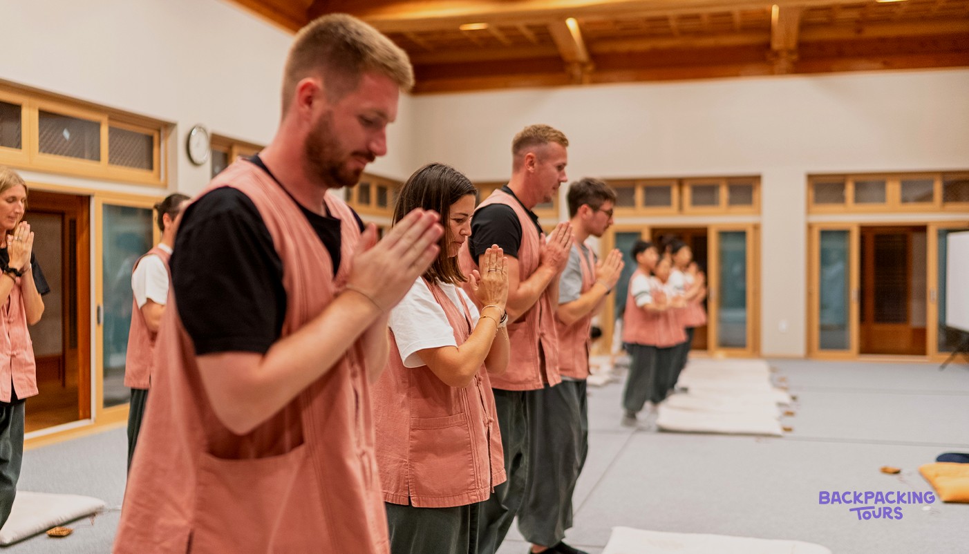 Travellers in temple-stay vests standing with hands pressed together during prayers at a Buddhist temple.