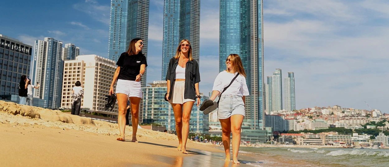 Three travellers walking along Haeundae Beach with the Busan skyline rising behind them.
