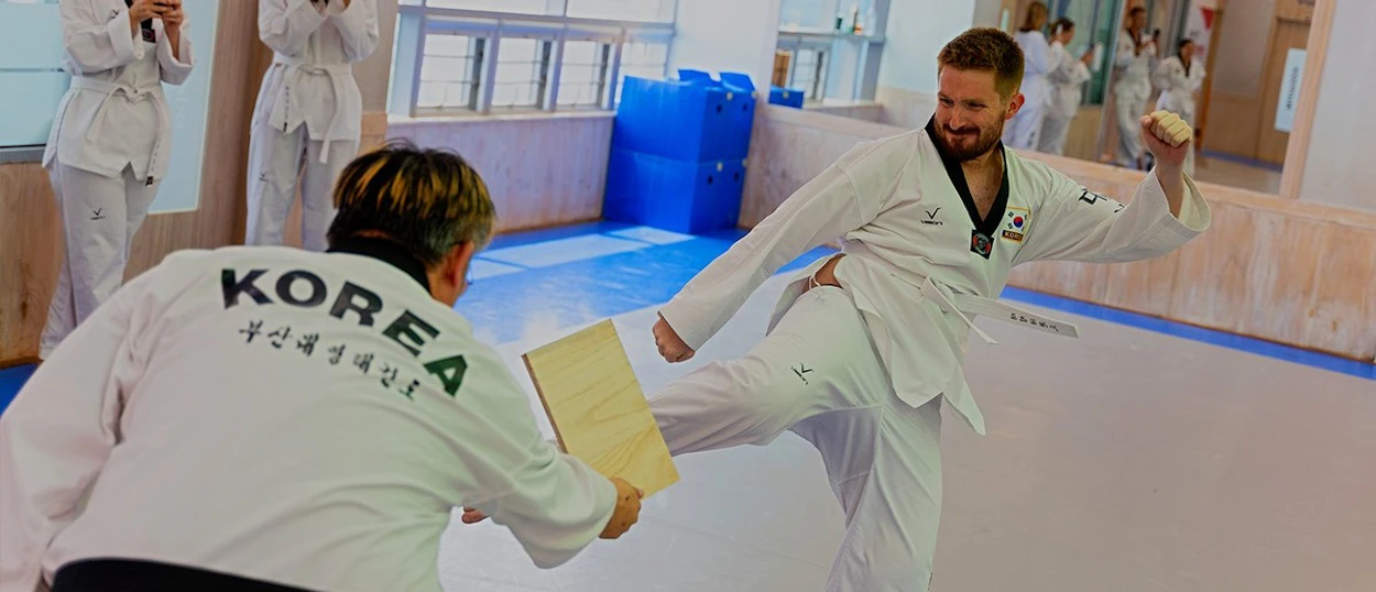 A taekwondo master holding a board while a student in a white dobok lands a flying kick.