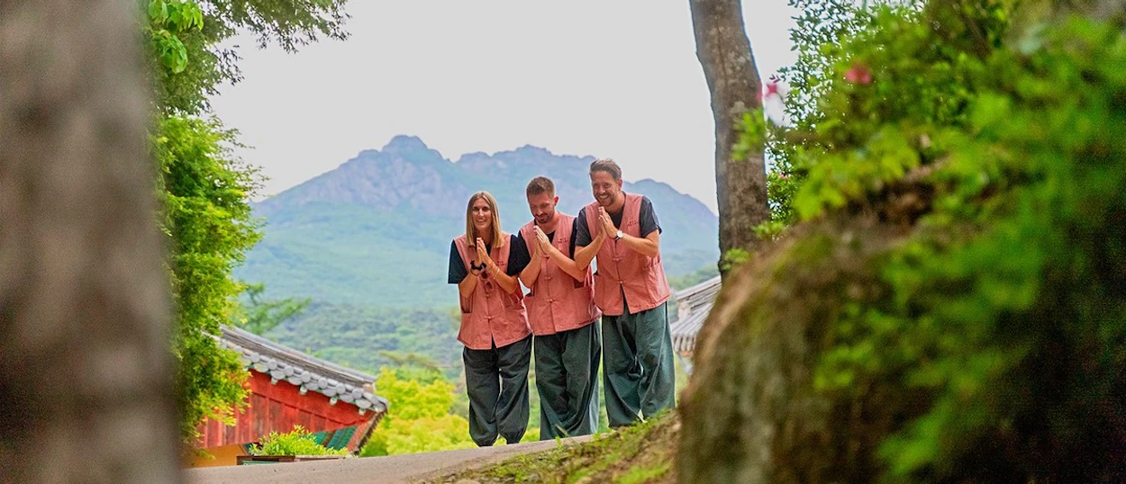 Travellers in pink temple-stay vests standing in front of the mountains at a Korean Buddhist temple.