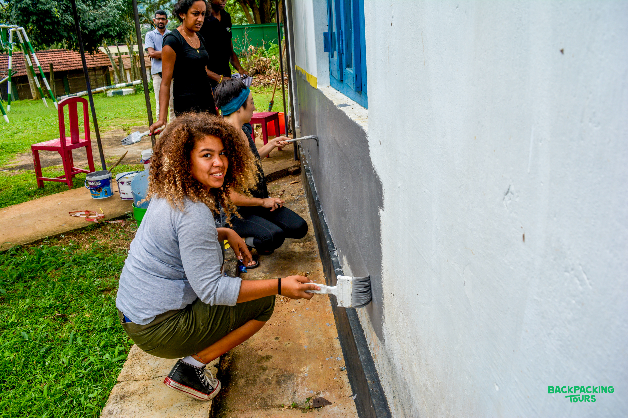 A smiling volunteer with curly hair paints a building wall grey during an outdoor renovation project.