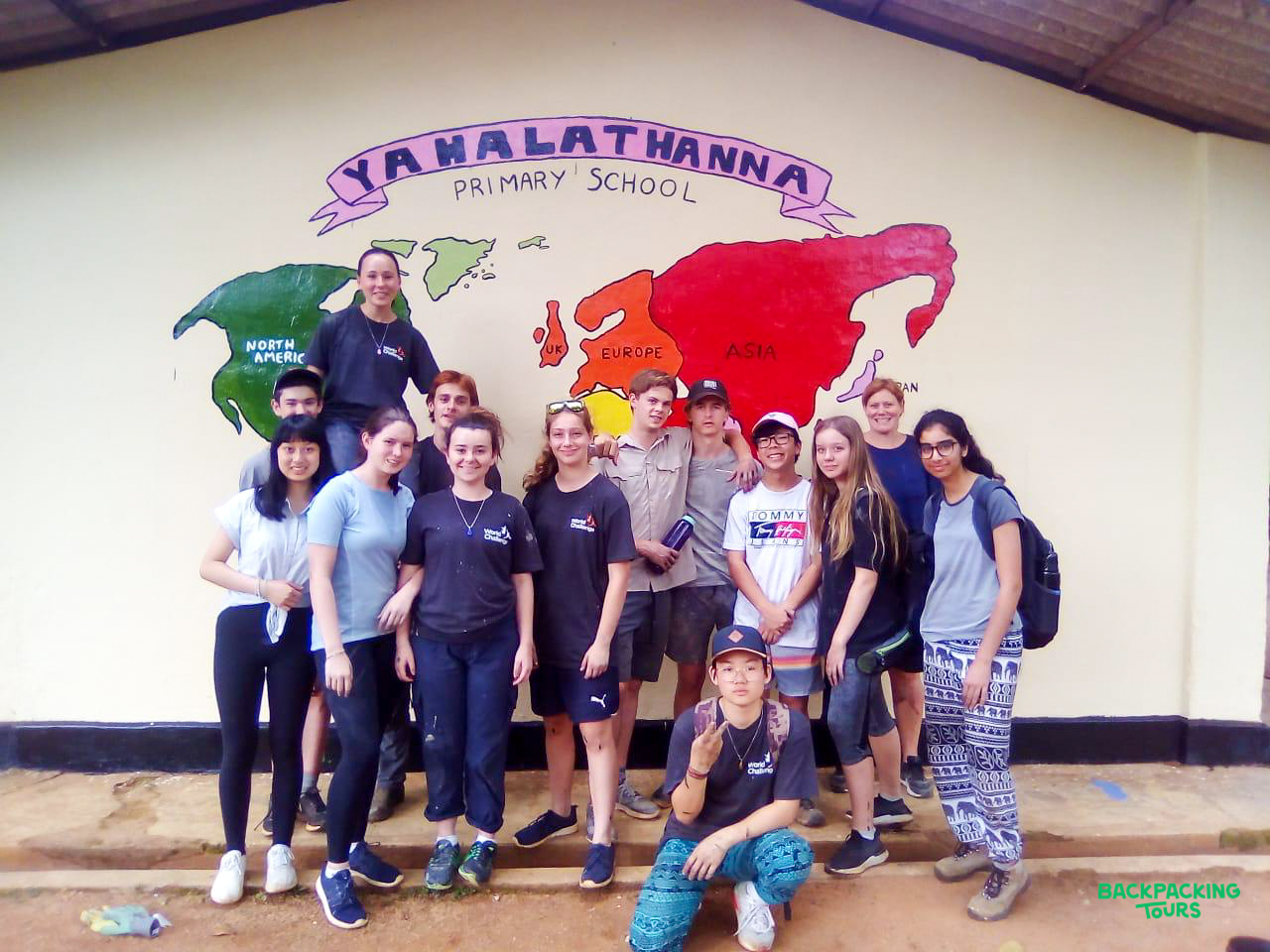 Volunteers pose happily in front of a colorful world map mural at Yahalathanna Primary School in Sri Lanka.