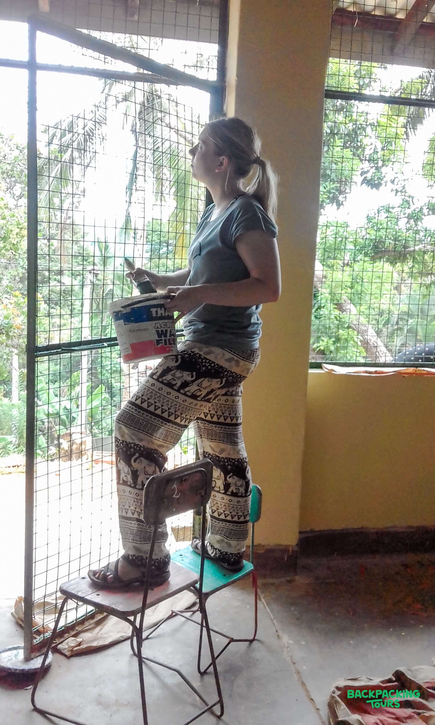 A woman stands on two chairs, painting a window frame with lush Sri Lankan greenery visible outside.