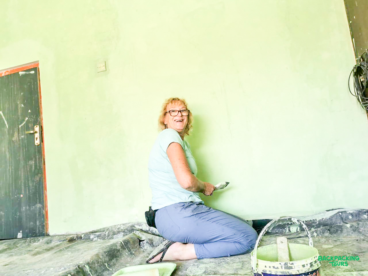 A smiling woman kneels, painting a light green wall during a renovation project in Sri Lanka.