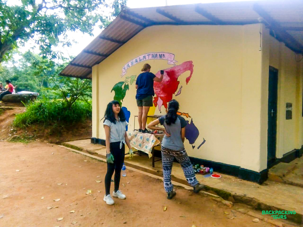 Three volunteers paint a colorful world map mural on a building in Sri Lanka's lush countryside.