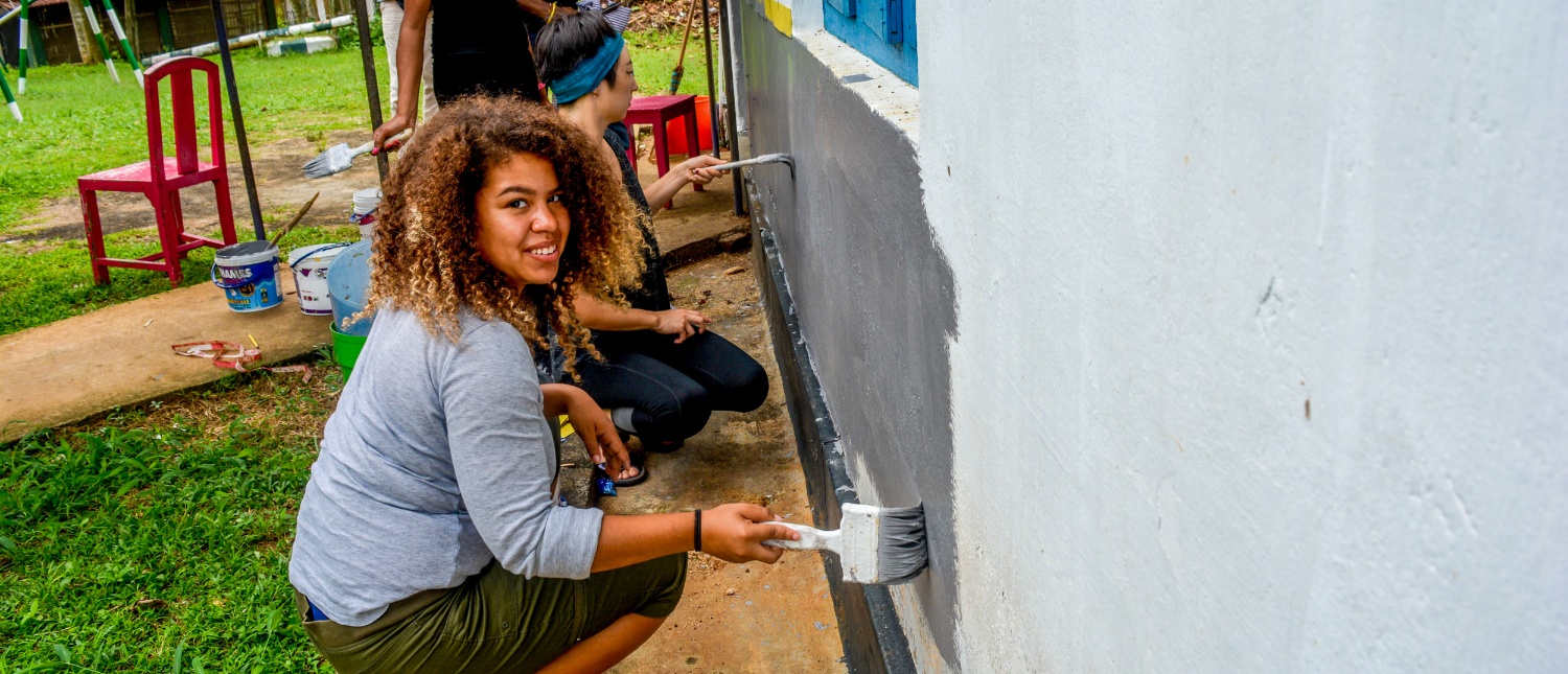 A smiling woman paints a building wall grey outdoors, contributing to a renovation project.