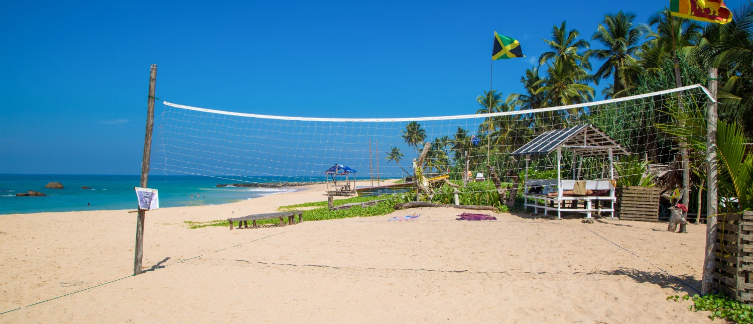 A tropical beach in Sri Lanka with a volleyball net, palm trees, and Sri Lankan and Jamaican flags.