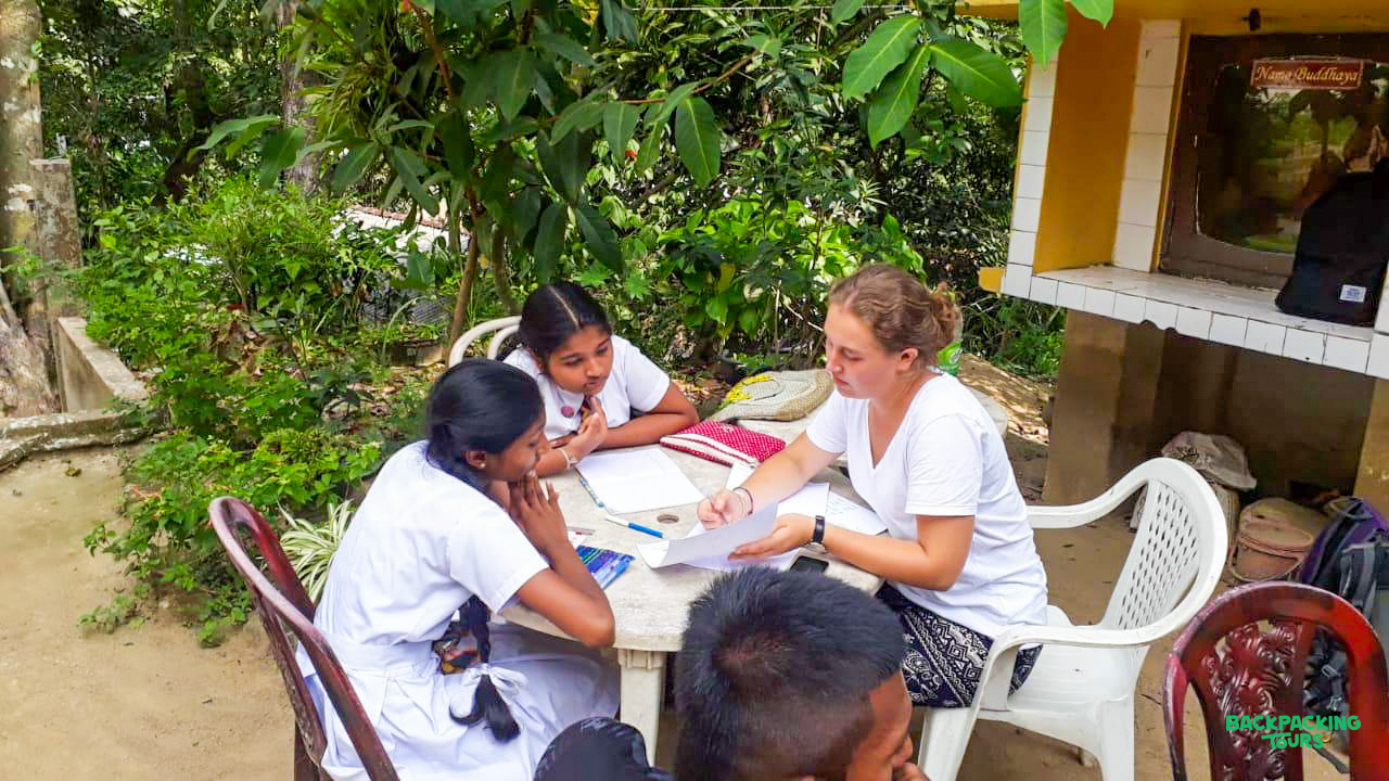 Volunteer teacher guides two local students at an outdoor table amidst Sri Lanka's lush greenery.