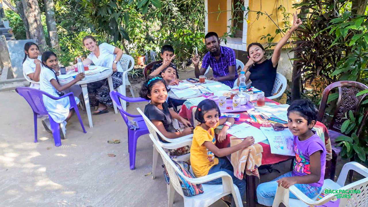 Smiling children and volunteers enjoy drawing and learning together at outdoor tables in Sri Lanka.