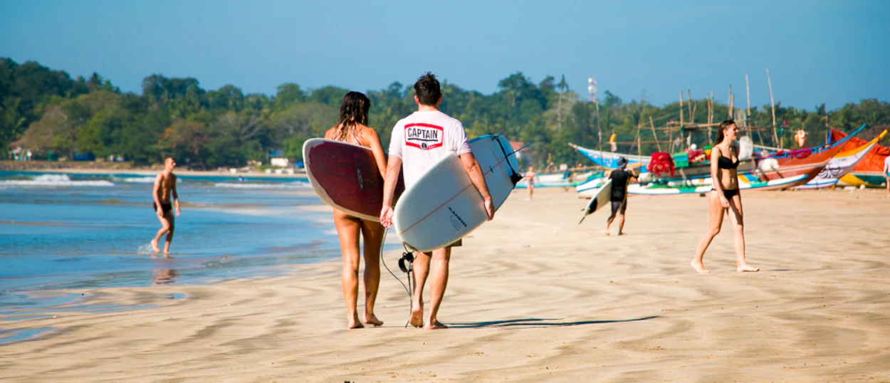 A man and woman carry surfboards along the sunny Weligama Beach in Sri Lanka.