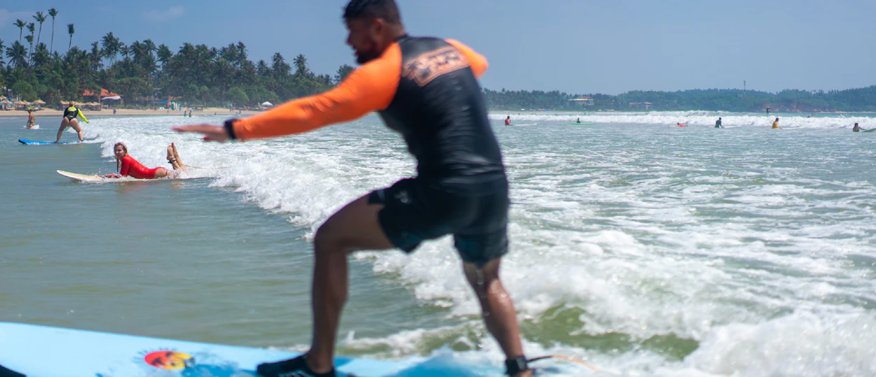 A man stands on a blue surfboard, learning to ride a wave at Weligama Beach.