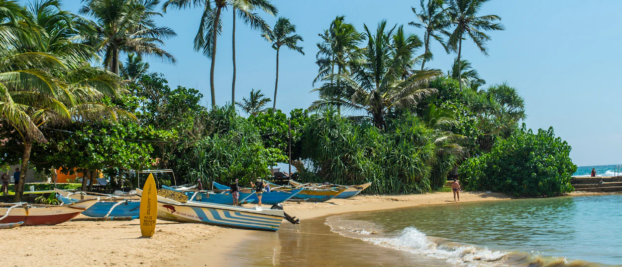 Traditional fishing boats and a surfboard on a sunny beach with palm trees in Mirissa, Sri Lanka.