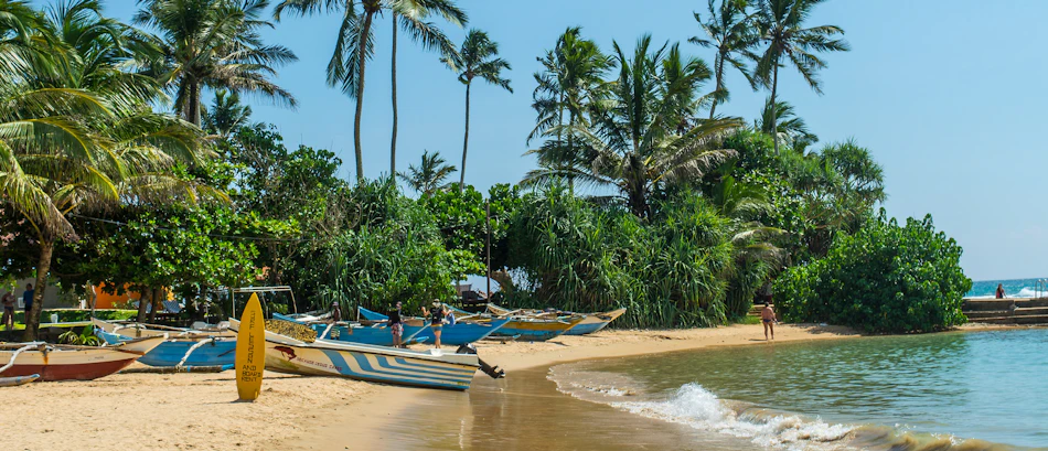 Traditional fishing boats and a yellow surfboard on a sunny tropical beach with palm trees in Unawatuna.