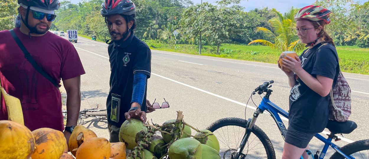 Two cyclists, wearing helmets, enjoy refreshing coconuts from a roadside stall in Unawatuna, Sri Lanka.