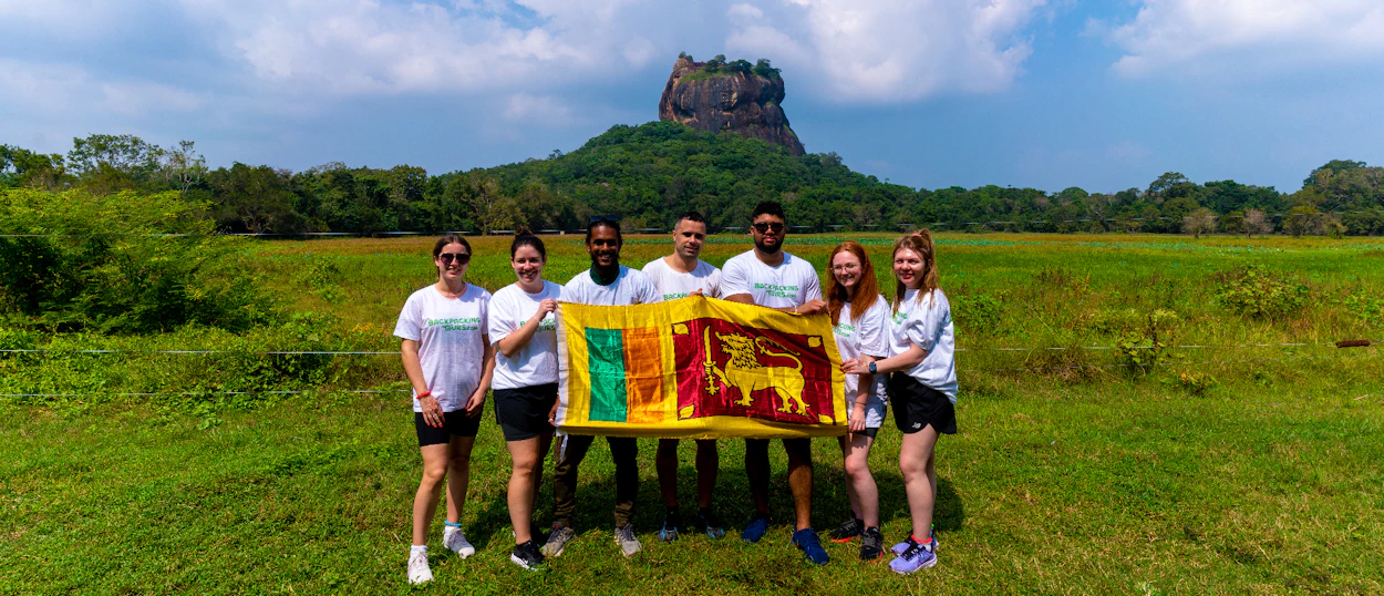 Six happy backpackers hold a Sri Lankan flag in front of Sigiriya Rock.