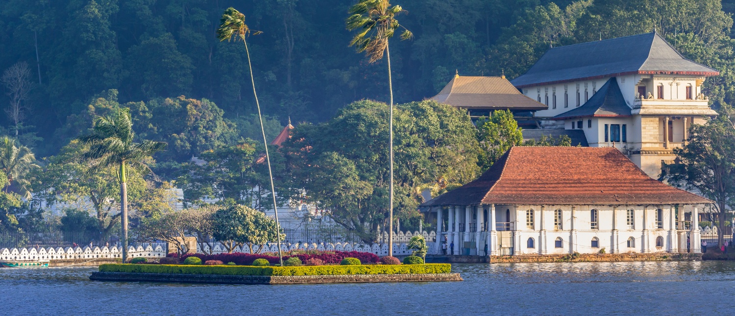 Historic buildings with red roofs and white columns on a lake, surrounded by lush green hills in Kandy, Sri Lanka.