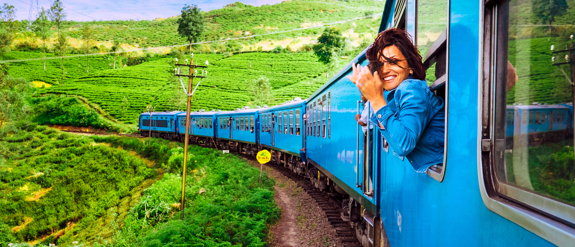 A joyful traveler leans from a blue train window, waving through green tea plantations in Sri Lanka.