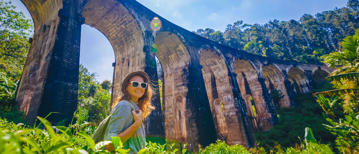 Happy traveler smiles in front of the historic Nine Arch Bridge amidst Sri Lanka's green hills.