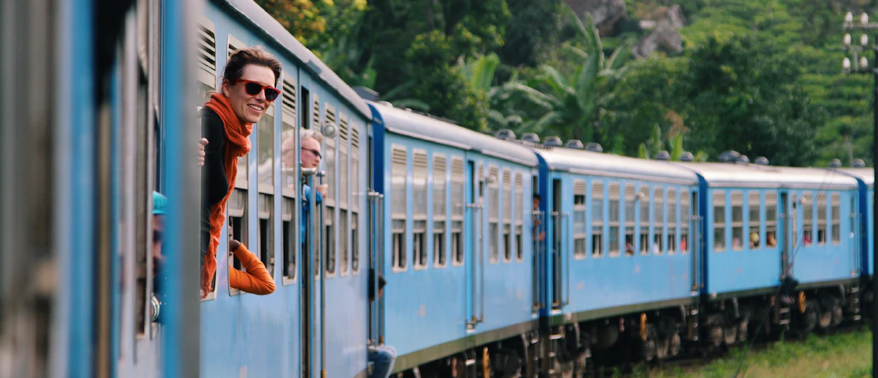 A smiling traveler leans out of a blue train window, enjoying the lush Sri Lankan landscape.