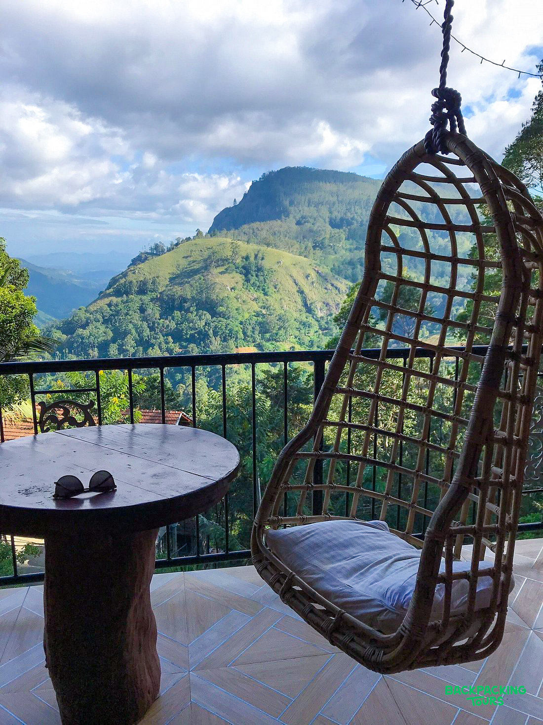 A cozy balcony with a wicker hanging chair and stone table offers stunning views of Ella's green mountains.